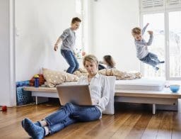 Anya laptopon dolgozik a földön, míg a gyerekek az ágyon ugrálnak.