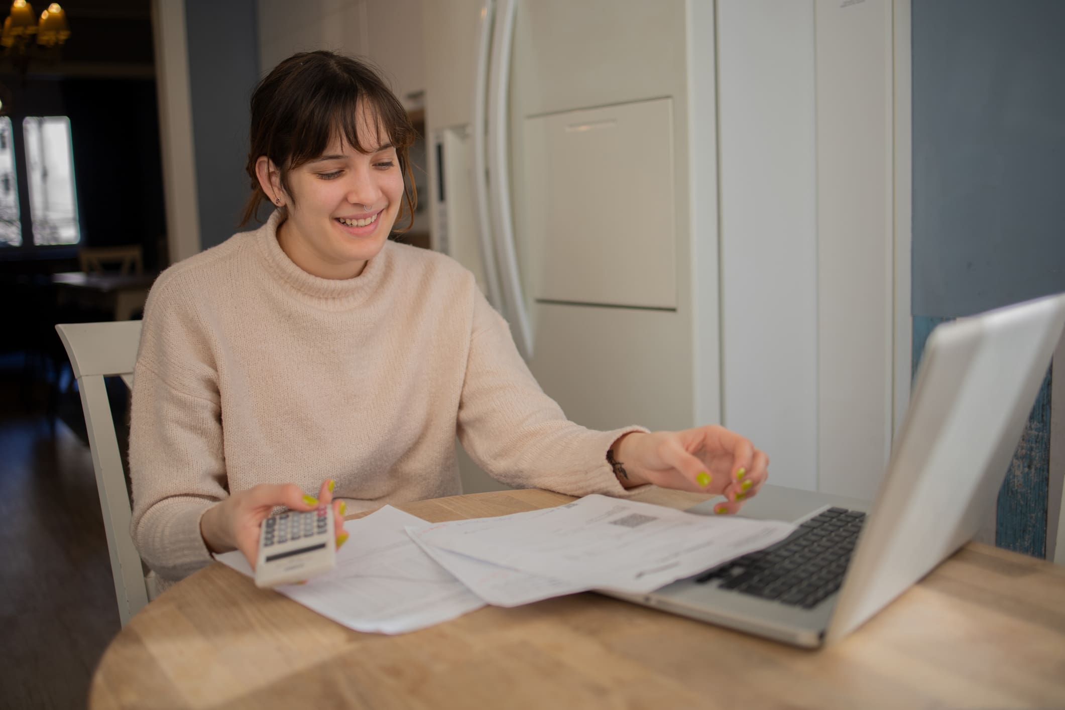 Egy fiatal nő kalkulál az előtte lévő laptopon, számológéppel, és papírokat néz.