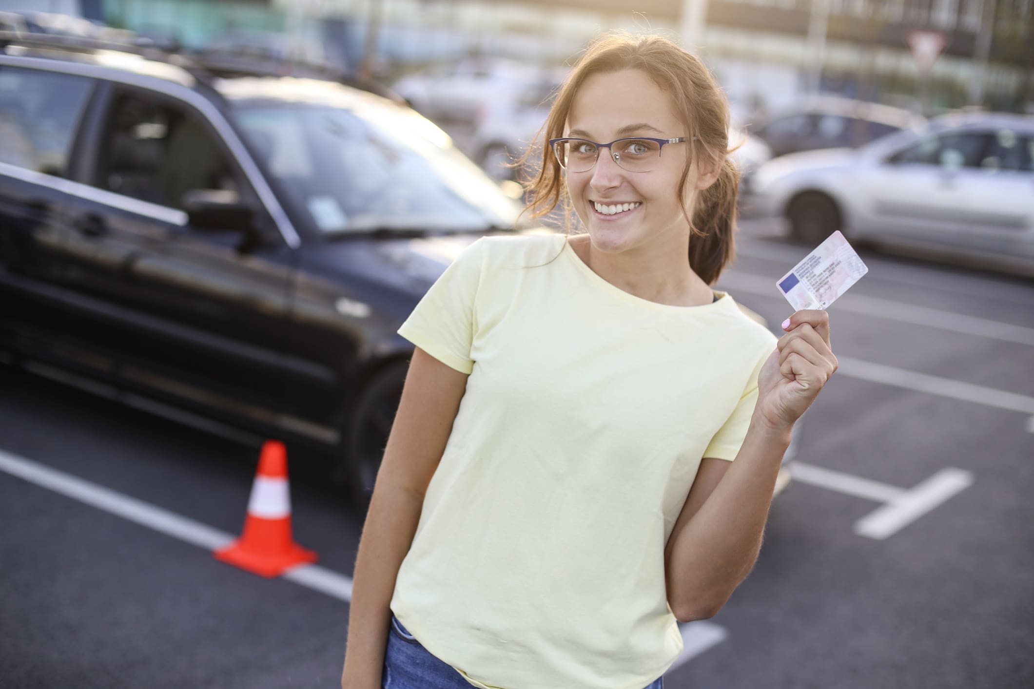 Egy fiatal lány mutatja fel az újonnan szerzett jögosítványát, háttérben egy fekete autó.