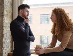 Szomorú fiatal pár az ablaknál, a férfi áll, a nő ül egy csészével a kezében.