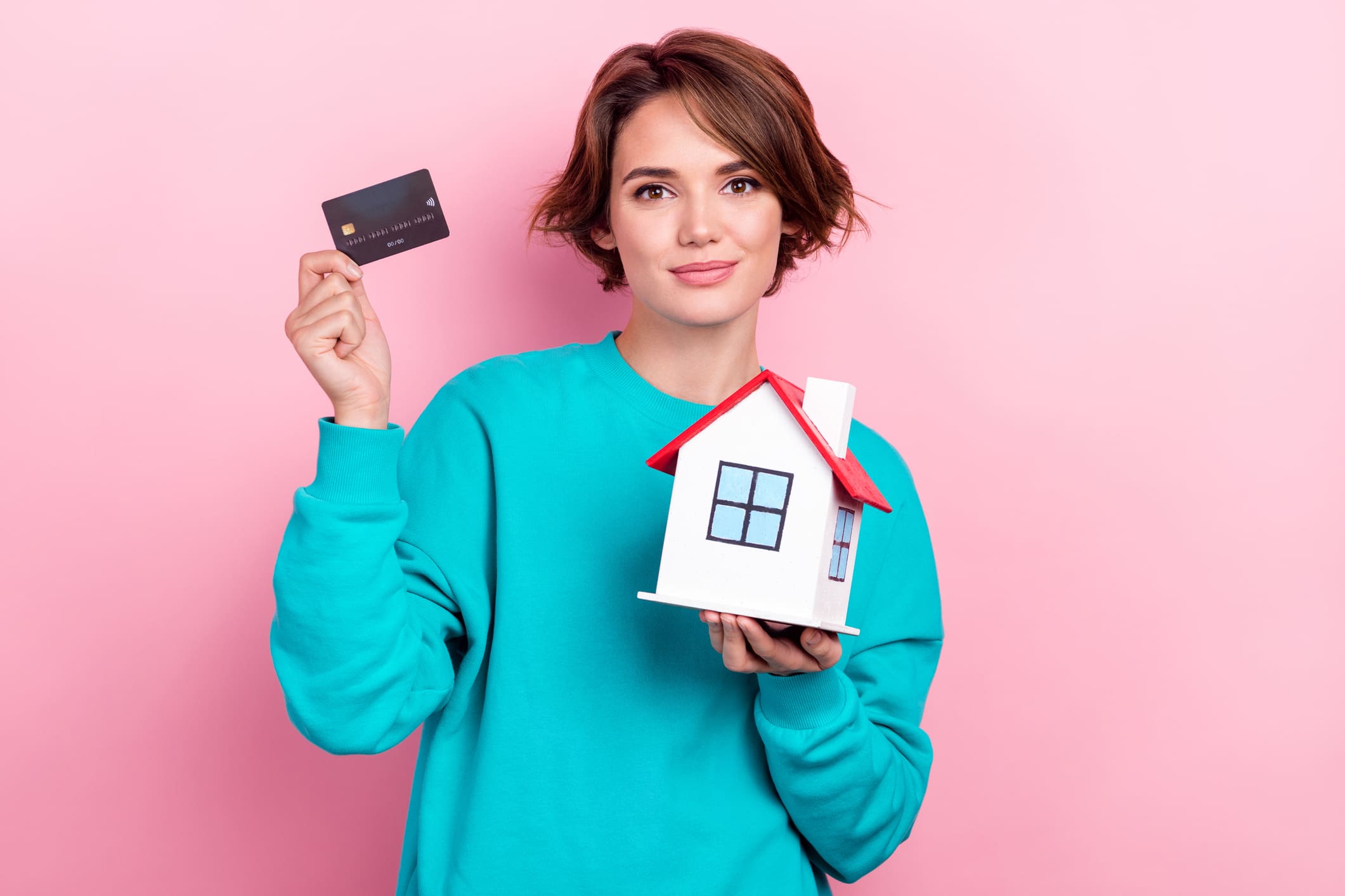 fiatal nő türkíz pulóverben, rózsaszín fal előtt, kezében bankkártya és egy házikó. 20 millió forintot utal át az új lakásáért