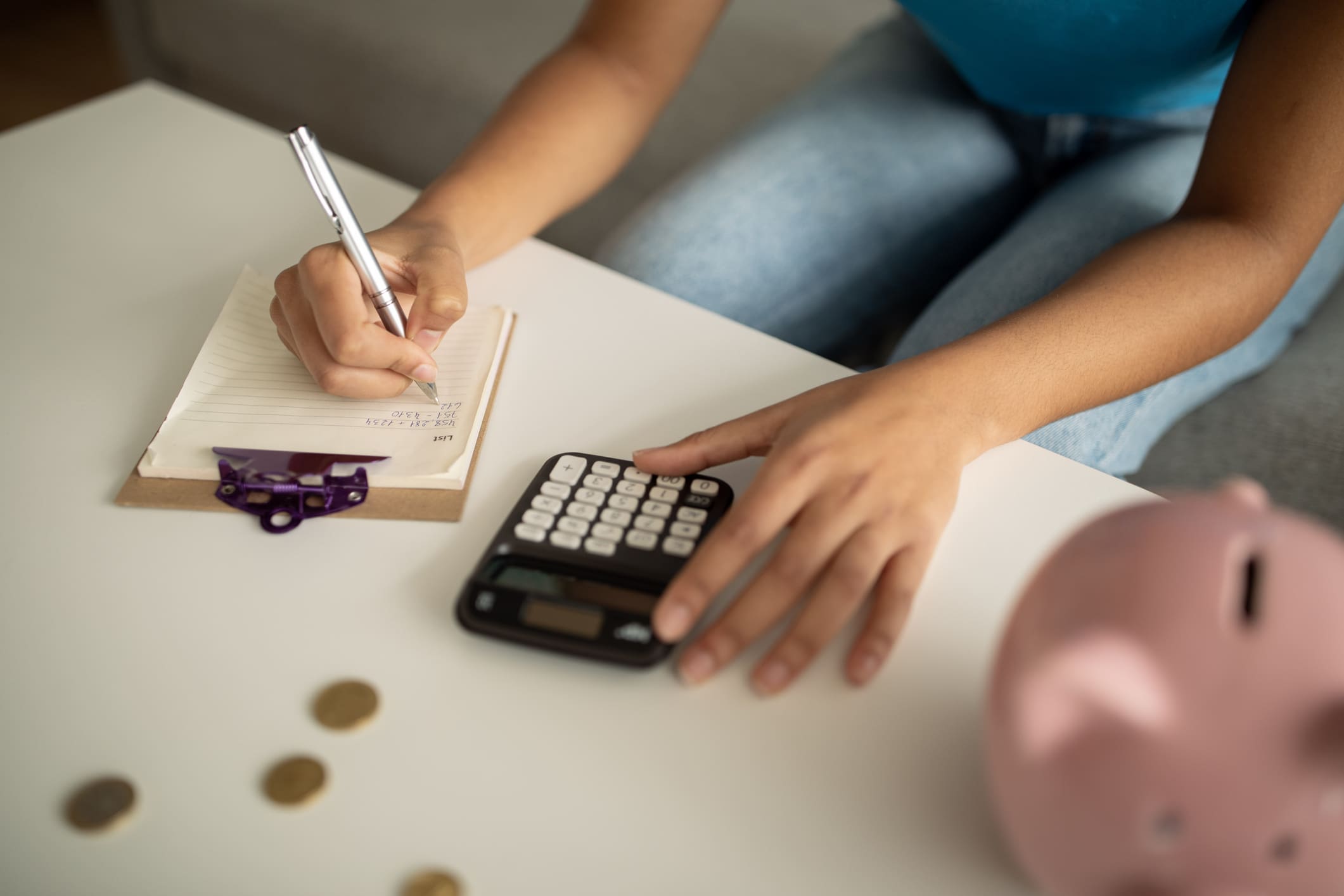 Íróasztalon malacpersely, pénzérmék, számológép, jegyzetfüzet, egy nő tollal jegyzetel, közben számol 