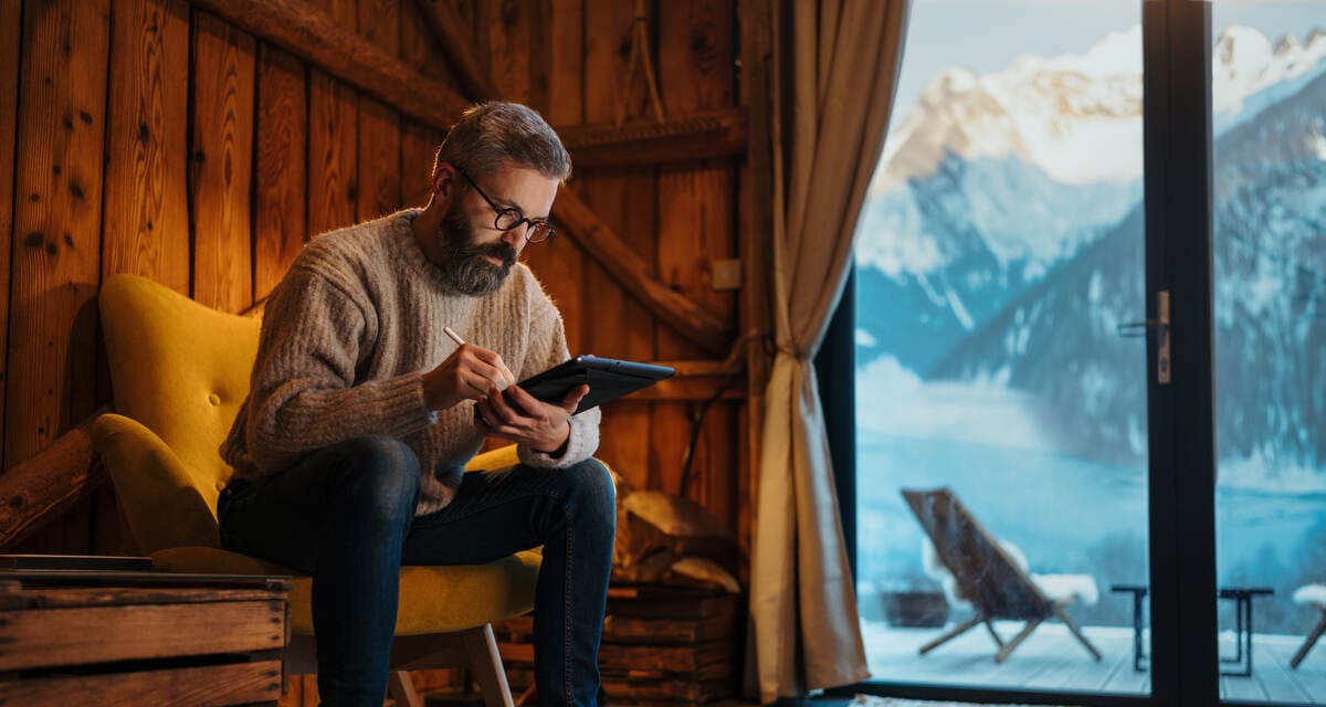 középkorú férfi faházban, odakint havas hegyek, kezében laptop, tél van, a nyaralására készül