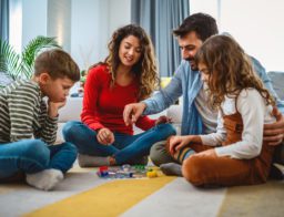 Egy család egy nagyonn fiú és egy kisebb lánygermekkel társasjátékoznak a nappali szőnyegén.