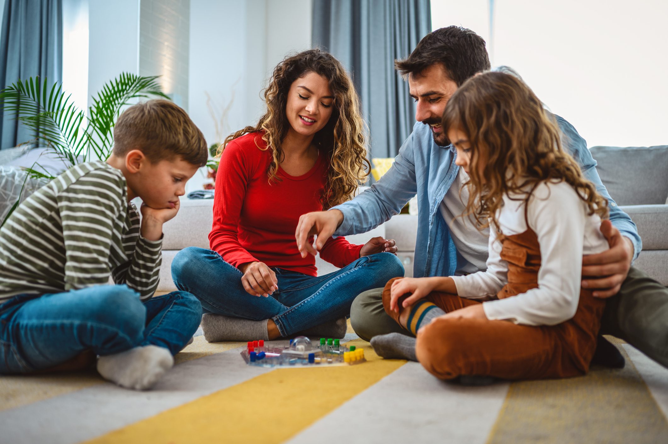 Egy család egy nagyonn fiú és egy kisebb lánygermekkel társasjátékoznak a nappali szőnyegén.