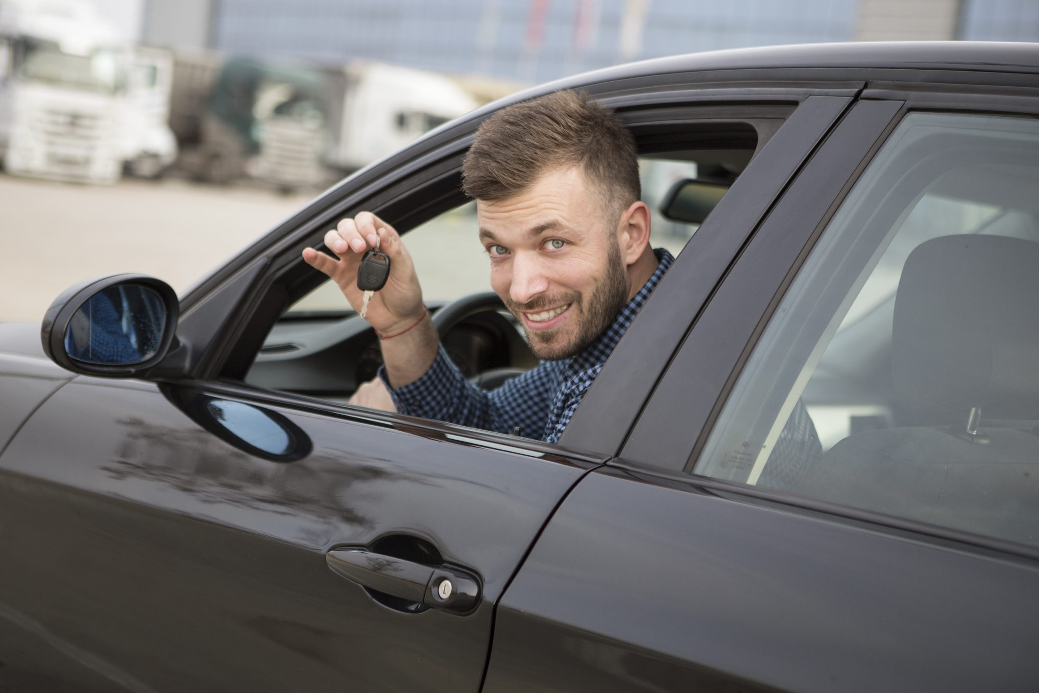 Egy fiatal férfi ül az új autójában és a lehúzott ablakon keresztül mutatja az új autó kulcsát.