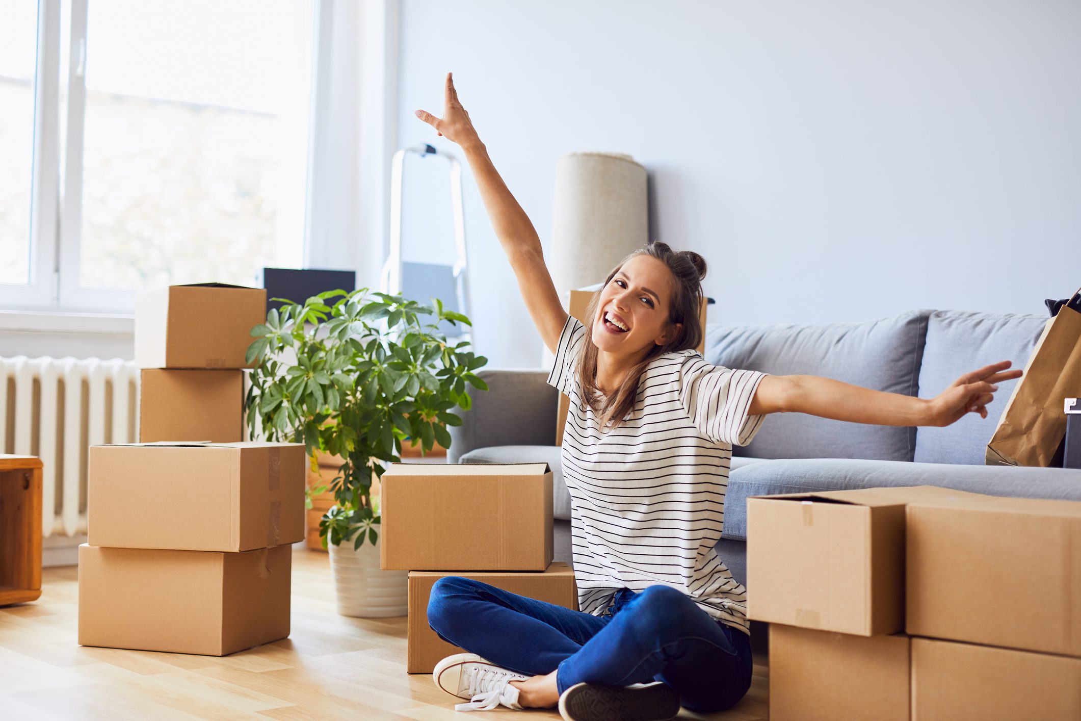 Fiatal nő a padlón ülve, magasba tartott kézzel örül az új lakásba költözésnek. Körülötte költözős dobozok.