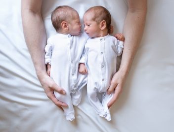 Iker babák fehér rugdalózóban fekszenek egy ágyon anyjuk ölelő karjaiban.