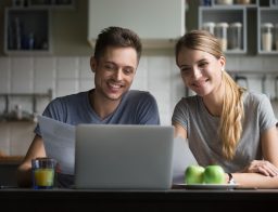 Egy fiatal pár boldogan ül egy laptop előtt és a férfi kezében egy dokumentum is van