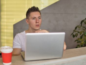 Egy fiatal fiú csodálkozva ül egy laptop mögött, melleteaz asztalon fedeles papírpohár.