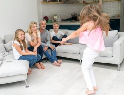 Háromgyermekes család játszik a nappaliban: két szülő, két gyerek ülnek a kanapén, egy kislány ugrál