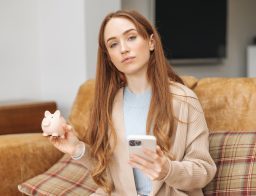 Egy hossú, vörös hajú nő ül egy kanapén, egyik kezében mobiltelefon, másikban malacpersely.