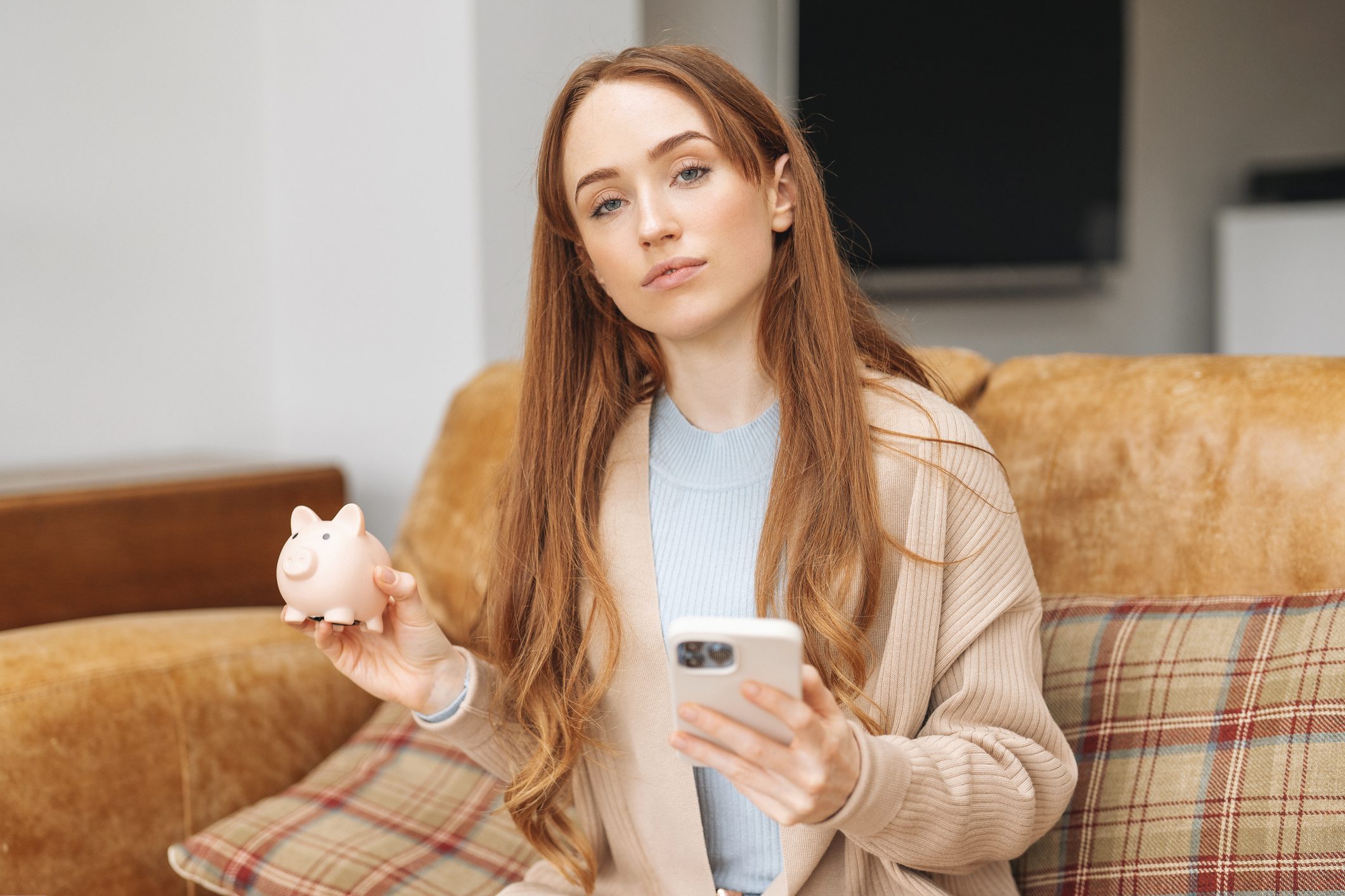 Egy hossú, vörös hajú nő ül egy kanapén, egyik kezében mobiltelefon, másikban malacpersely.