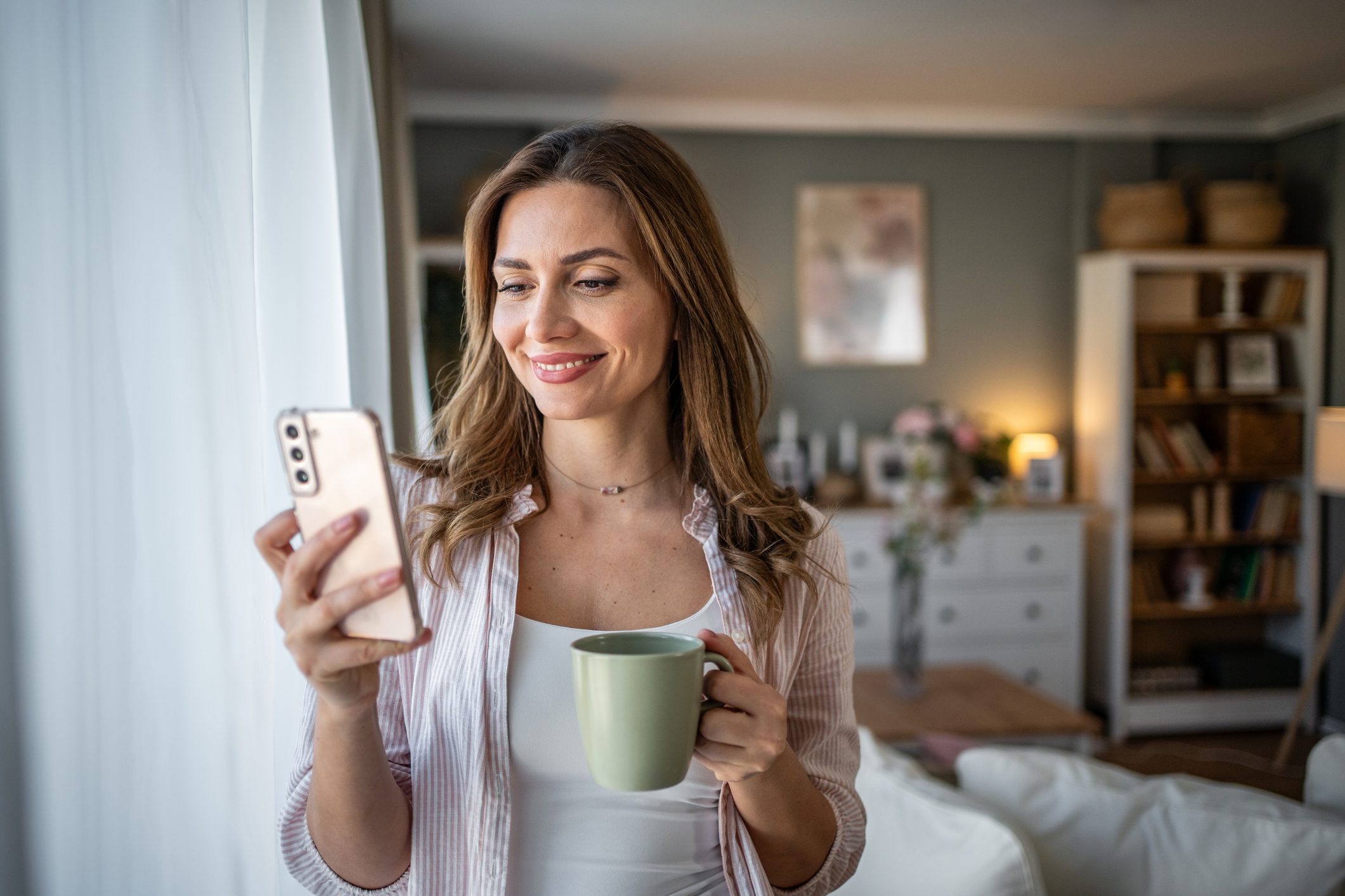 Kozépkorú nő otthon, kezében kávéval, mobiltelefonnal
