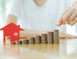 Egy fehér pólós nő eegy piros házikó ikon mellett érméket pakol egymásra oszloponként lépcsőzetesen.