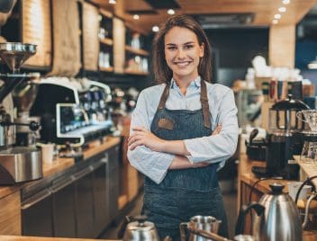 Egy fiatal, hosszú barna hajú lány világoskék ingben, szürke kötényben áll a kávézó pultjában