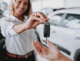 Egy nő átadja az autókulcsot valakinek, akinek csak a kezét látni. Háttérben egy fehér autó.
