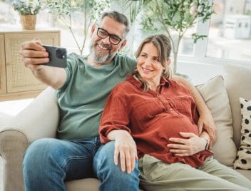 Idősebb házaspár gyermekvállalás. Kanapén ülnek, a férfi selfie-t készít a telefonjával.