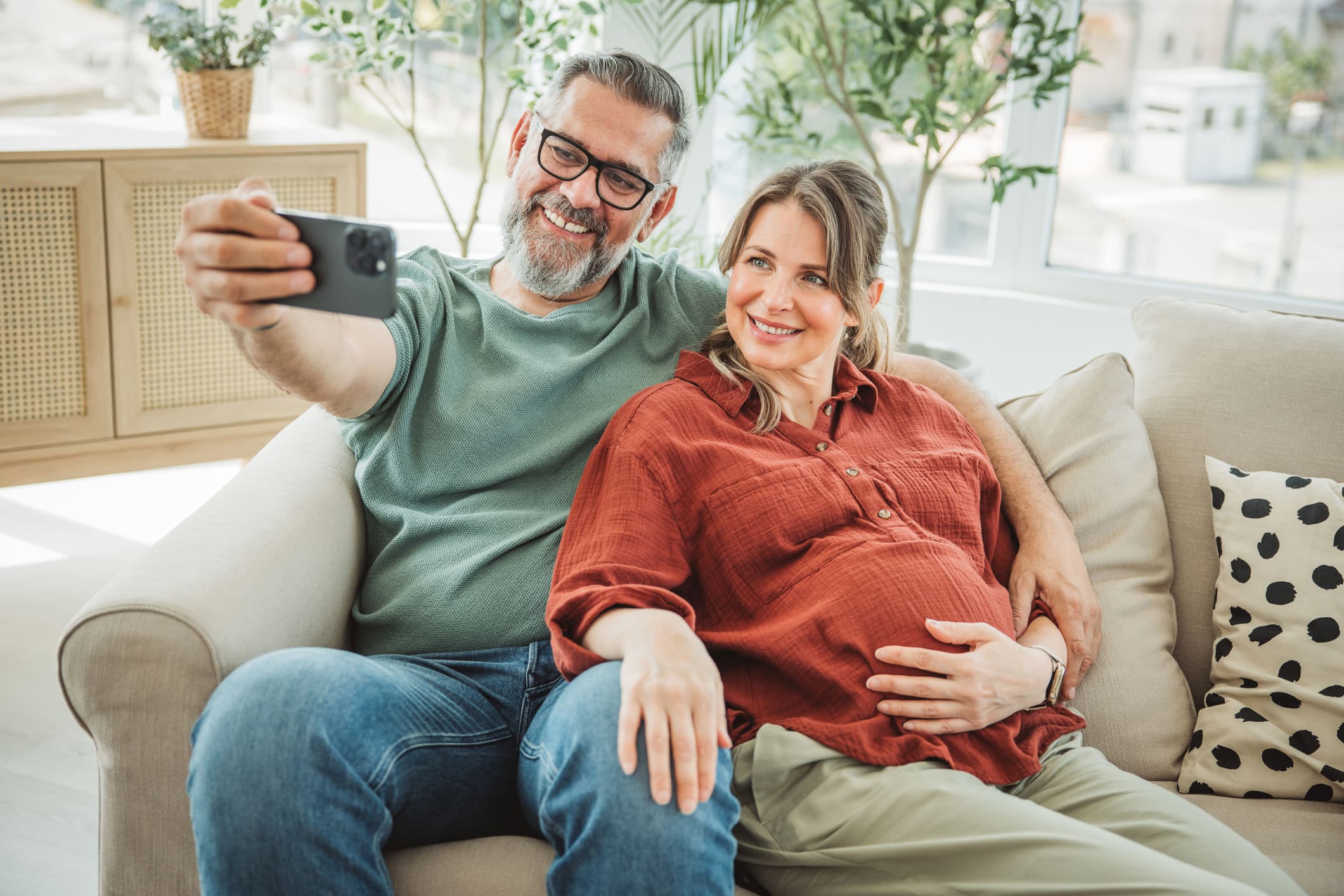 Idősebb házaspár gyermekvállalás. Kanapén ülnek, a férfi selfie-t készít a telefonjával.