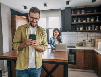 Egy pár férfi tagja a konyhasziget előtt áll és a mobilját nézi, a nő pedig a konyhaszigat mögött állva a laptopot nézi.