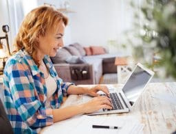 Egy középkorú nő otthoni ruhában üle egy laptop előtt, mellette az asztalon toll, papír.