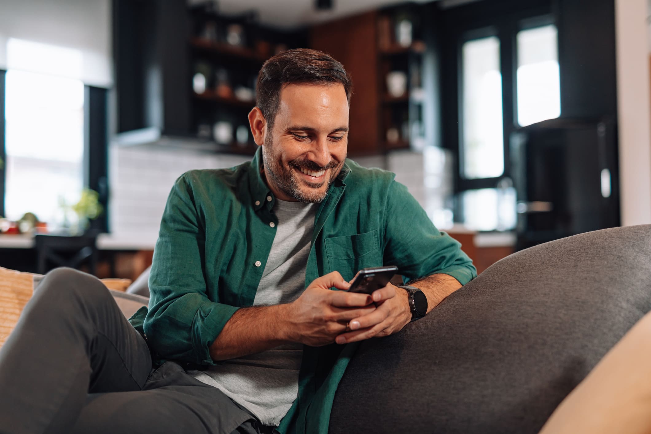 Egy zöld inges férfi nézni mosolyogva a mobiltelefonját a háza kanapéján.