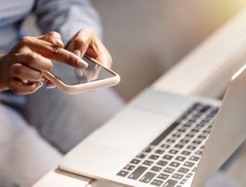 Egy női kéz az íróasztal mellett egyszerre használ mobiltelefont és laptopot.