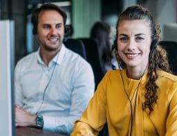 Call centeres dolgozók. A kép előterében egy fiatal nő sárga felsőben, mögötte egy fehér inges férfi ül, fülhallgatóval a fejükön.