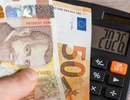 Egy emberi kézben egy bolgár leva és egy eurós bankjegy, mögötte számológép, aminek a kijelzőjén 2026 szerepel, utalva Bulgária euroövezeti csatlakozására.