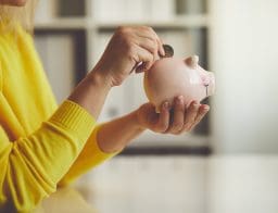 Egy sárga pulóveres női kéz beledob egy érmét egy rózsaszín malacperselybe, amit a másik kezében tart.