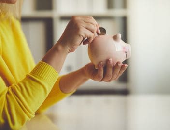Egy sárga pulóveres női kéz beledob egy érmét egy rózsaszín malacperselybe, amit a másik kezében tart.