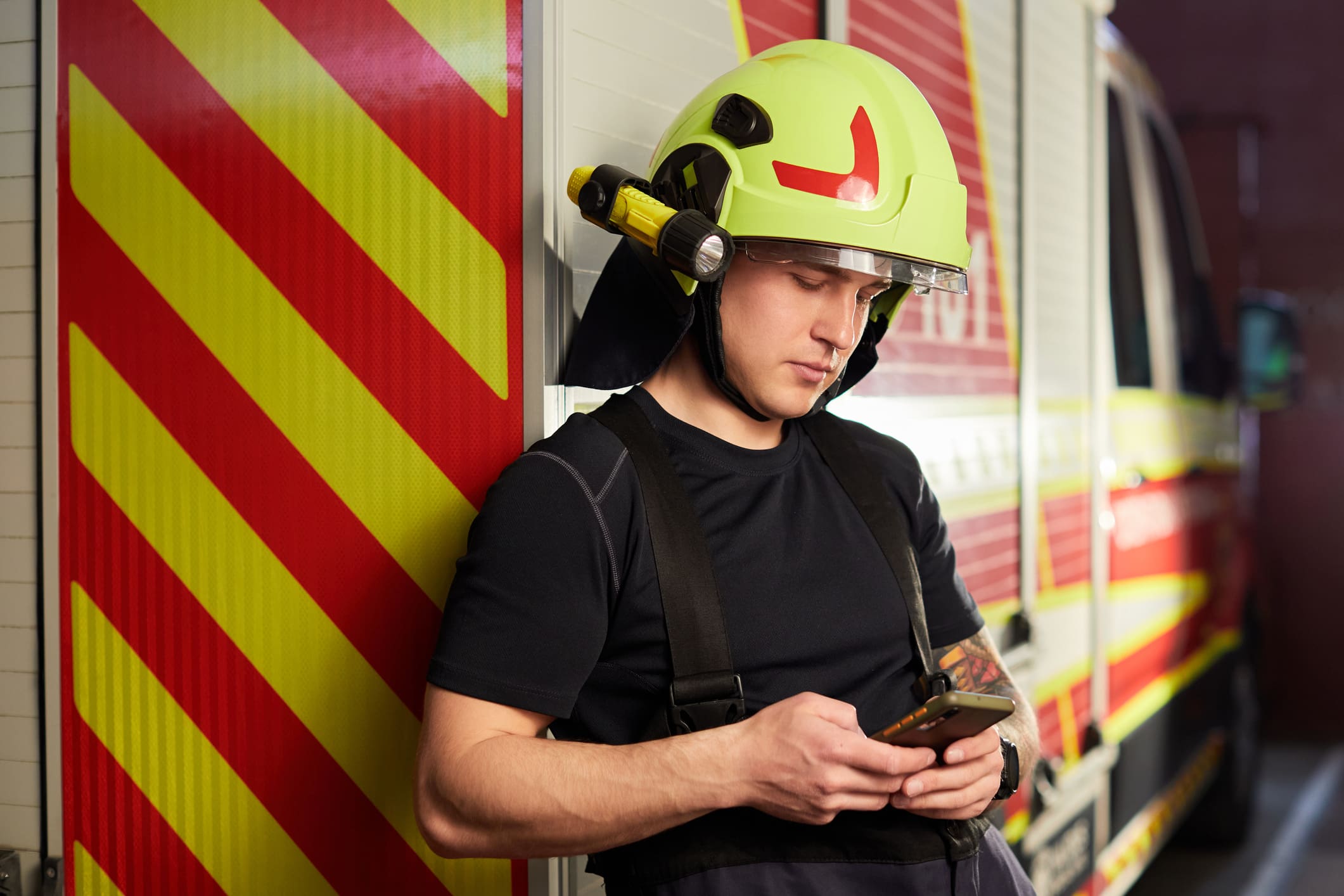 Tűzoltó háttal a tűzoltóautónak dőlve mobiltelefonozik.