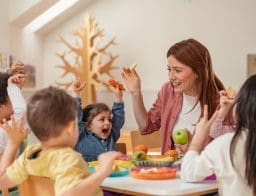 Bölcsödei uzsonna, gondozónő és négy gyermek látható egy kis asztalnál ülve, előttük gyümölcsök