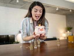 Egy ujjongó fiatal nő tart a kezében egy fehér-rózsaszín malacperselyt, az ebédlőasztal felett. Az asztalon pénzérmék.