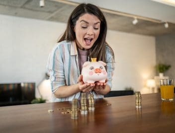 Egy ujjongó fiatal nő tart a kezében egy fehér-rózsaszín malacperselyt, az ebédlőasztal felett. Az asztalon pénzérmék.
