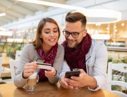 Fiatal pár egy kávézóban érdeklődve nézik a férfi mobiltelefonját, a nő kezében bankkártya.