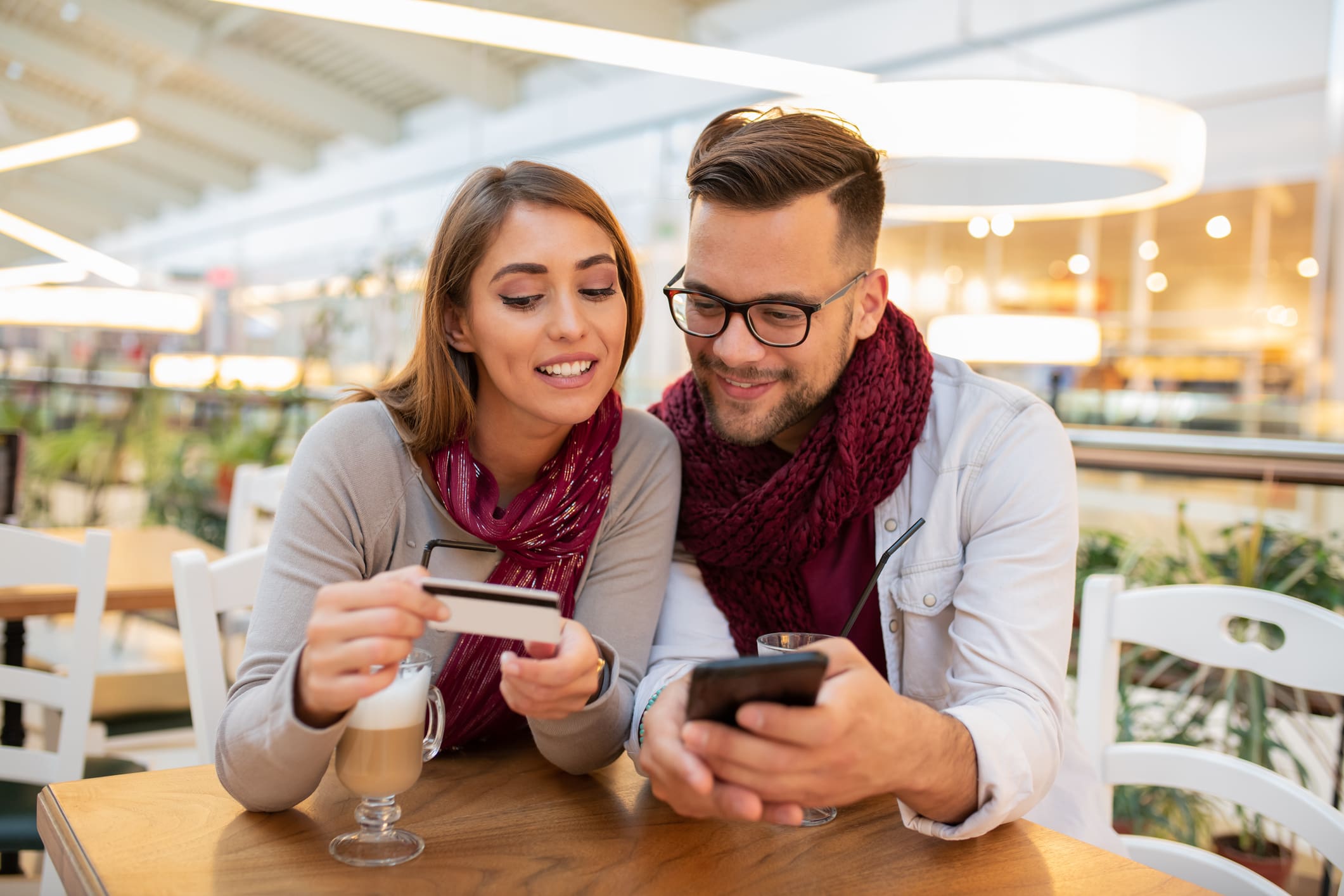 Fiatal pár egy kávézóban érdeklődve nézik a férfi mobiltelefonját, a nő kezében bankkártya.