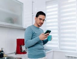 Egy fiatal szemüveges férfi a konyhában issza a kávéját, másik kezében a mobiltelefonja, arra figyel.