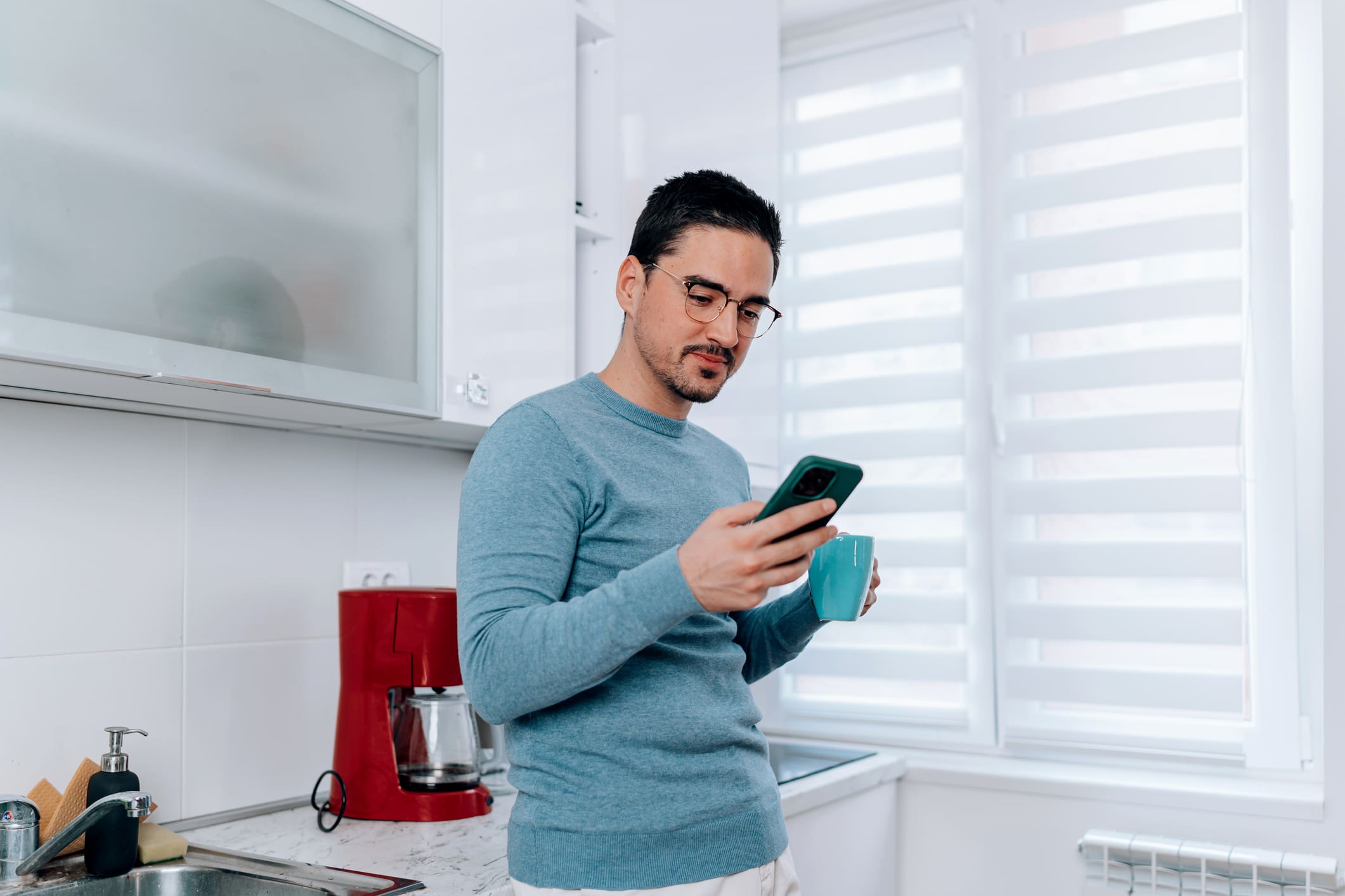 Egy fiatal szemüveges férfi a konyhában issza a kávéját, másik kezében a mobiltelefonja, arra figyel.