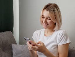 Egy fiatal szőke, rövid hajú lány otthon mosolyogva nézi a mobiltelefonját.