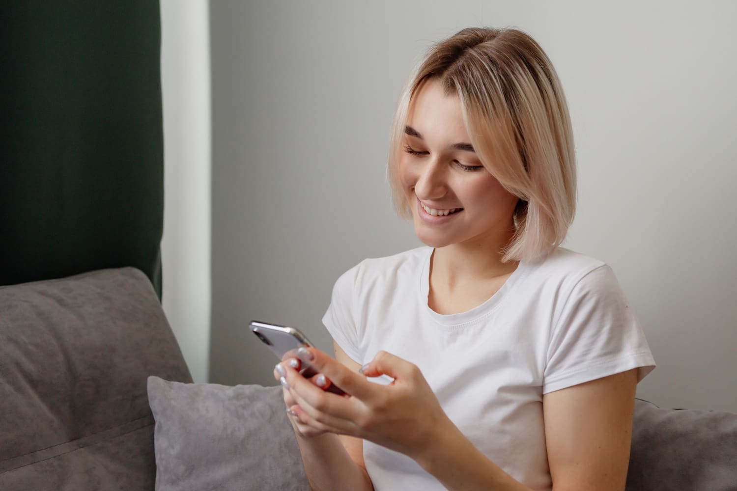 Egy fiatal szőke, rövid hajú lány otthon mosolyogva nézi a mobiltelefonját.