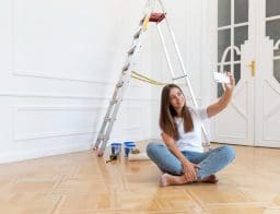 Egy barna hajú fiatal nő ül a felújított lakása padlóján, és szelfit készít. Háttérben egy étra és festékes vödrök.