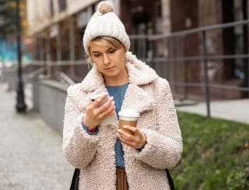 Egy nő nézi a telefonját az utcán, kezében papírpohárral. Kabátot, sapkát visel, intézmény kerítése előtt áll a járdán.