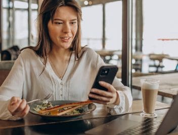 Fiatal vállalkozó hölgy ebédidőben nézi a mobiltelefonját, előtte laptop, tányér étel, tejeskávé,