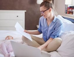 Egy fiatal anya ül az ágyon, előtte laptop és papír a kezében. Kisbaba mellette alszik.