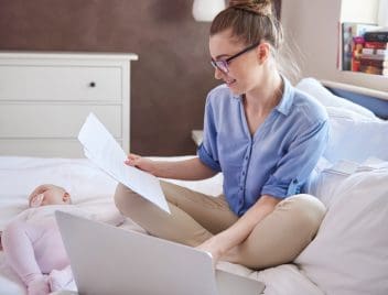 Egy fiatal anya ül az ágyon, előtte laptop és papír a kezében. Kisbaba mellette alszik.