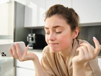 Egy fiata nő a konyhájában egyik kezében a telefonját fogva, kezeit széttárva csodálkozik. Elkedvetlenedett arcot vág.