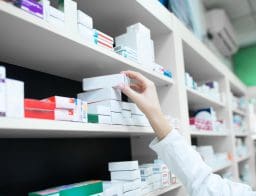 Gyógyszerész egy doboz gyógyszert vesz le a polcról egy gyógyszertárban, csak a keze és a polc látszik.