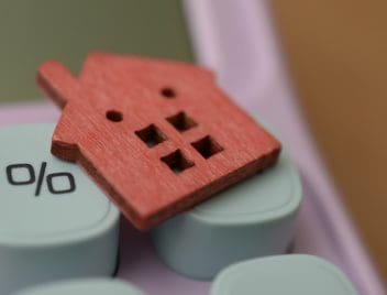 Egy részlet egy számológép nyomógombjairól, amelyek közül az egyiken százalékjel van. A számológépen pirosra festett kis házikó fából.