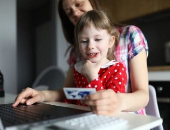 Egy fiatal anyuka ül egy laptop előtt, ölében a kislányával, kezében egy brosúrával.